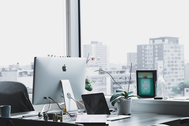 Modern office desk with iMac, laptop, orchid, and city view through a window